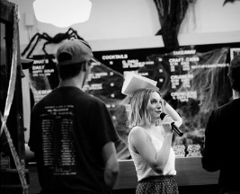Brydie on mic in front of a venue board, black and white